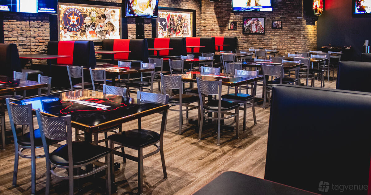 A bar with sports memorabilia, brick walls, booth and metal chair seating, and TVs on the walls at Texas Huddle.