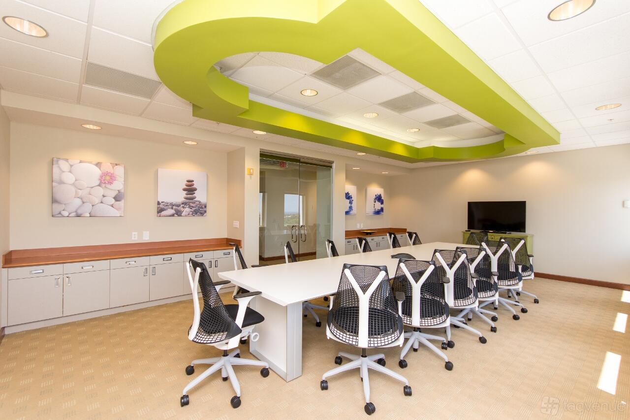 A boardroom with a long white conference table, ergonomic chairs, and green ceiling accents at ZEN Offices Boca Raton.