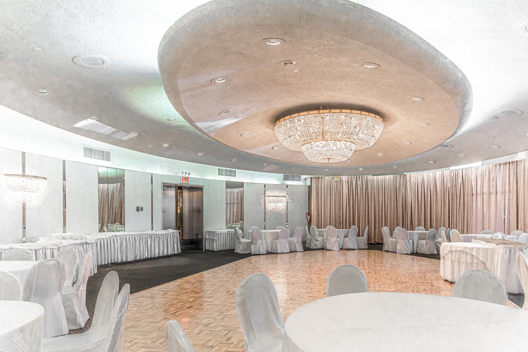 An event venue with a parquet dance floor, round tables covered in white linens, and a large crystal chandelier at Astoria World Manor.