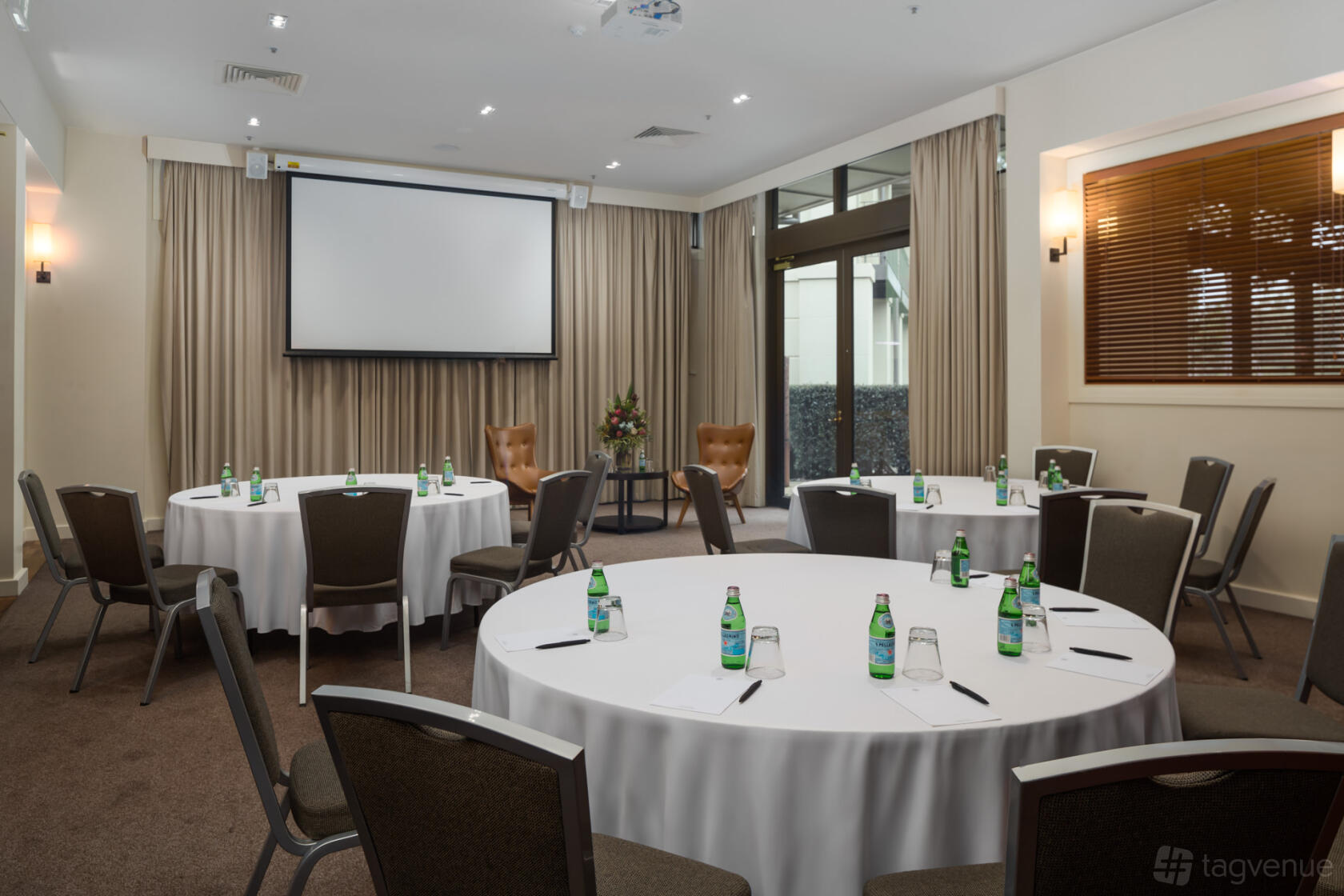 A hotel dining room with round tables dressed in white linens, bottled water, notepads, and a projector screen at Yarra Valley Lodge.