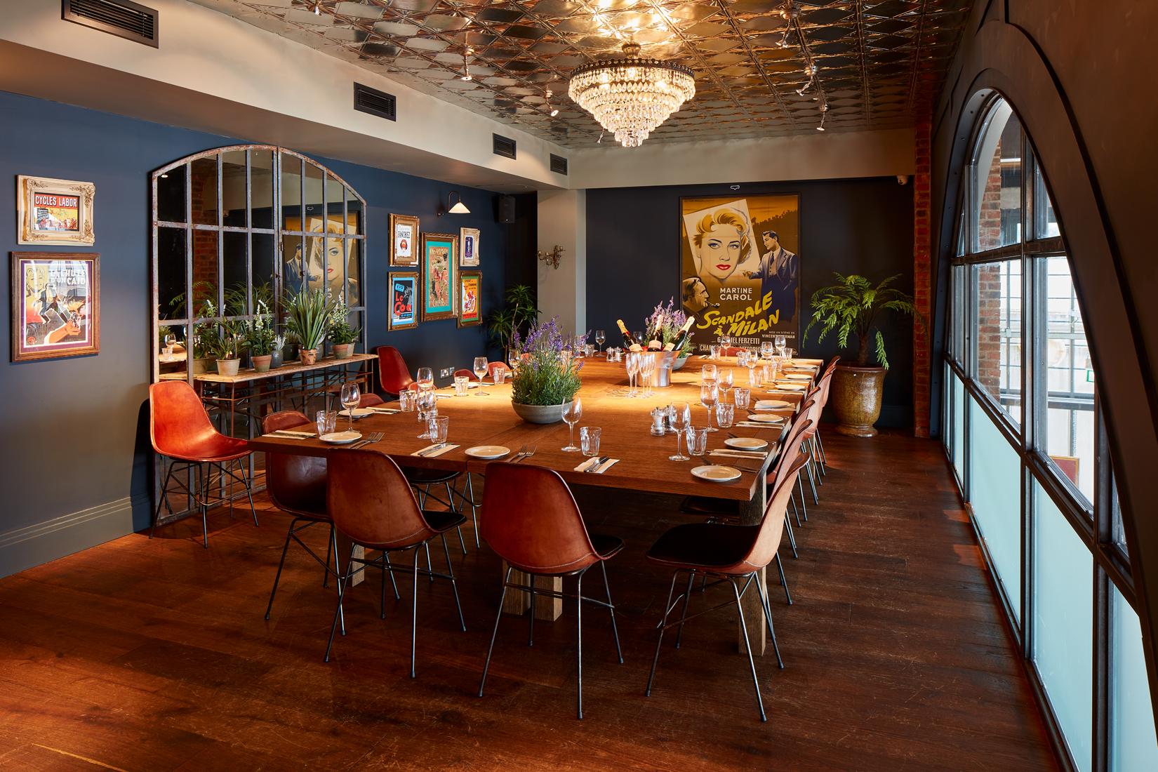 A dining room with a large wooden table, red chairs, chandelier, and framed art at Mumbles Bistrot Pierre.