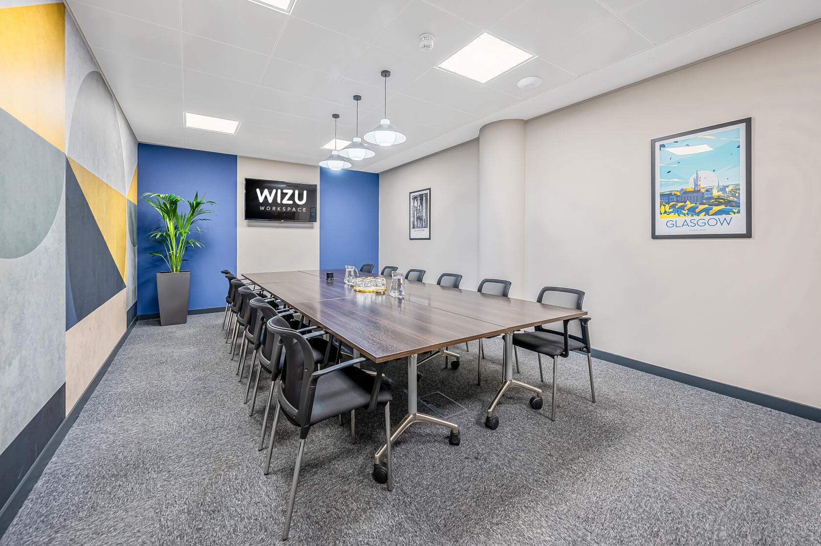 A meeting room with a long wooden table, black chairs, pendant lights, wall art, and carpeted floors at Wizu Workspace - Glasgow - West Regent Street.