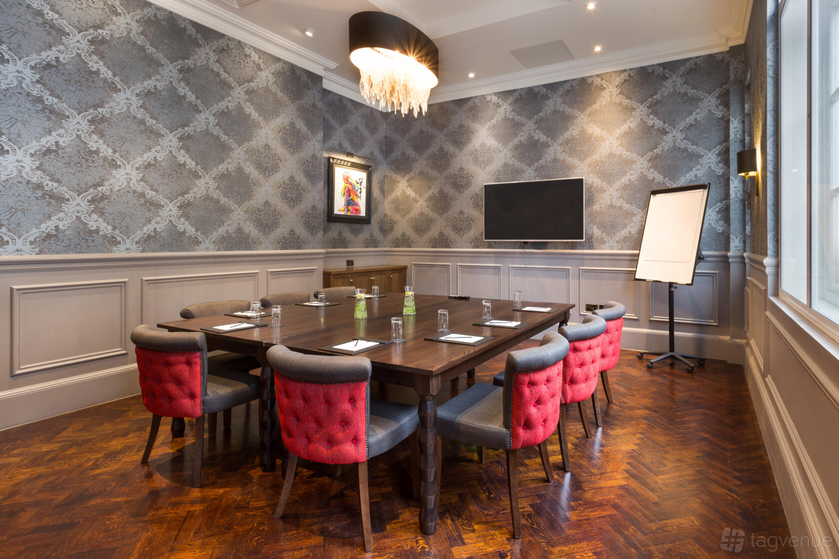 A private dining room with a wooden table, red cushioned chairs, chandelier, and wall-mounted TV at Courthouse Hotel Shoreditch.