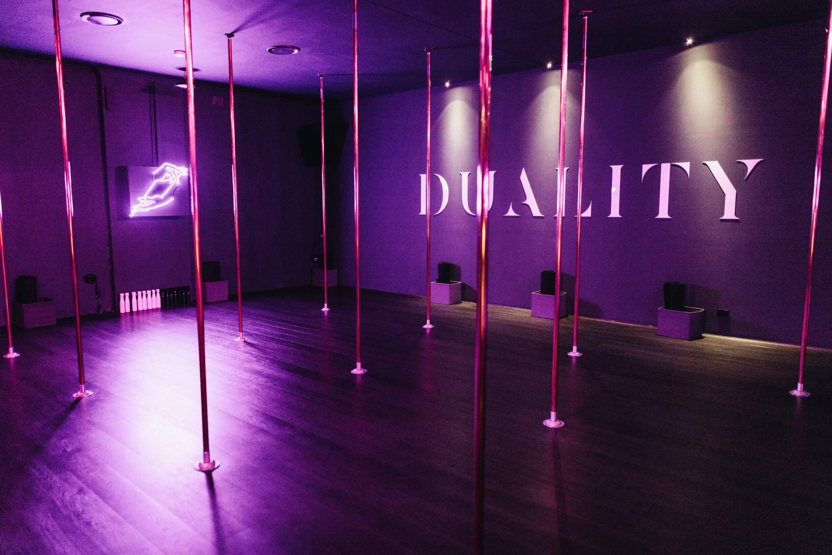 A dance studio with multiple pole stations, purple ambient lighting, and neon signage at Duality Pole Dancing Studio.