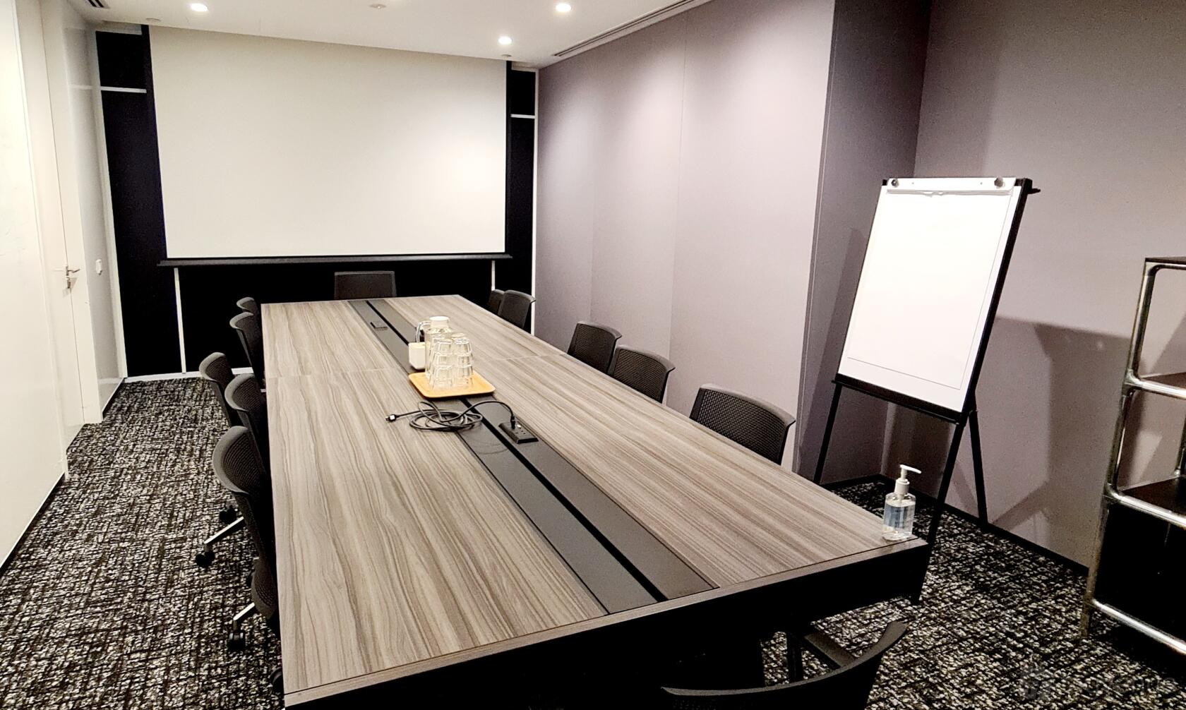 A meeting room with a long conference table, black chairs, projector screen, and flip chart at The Work Project OUE Downtown.