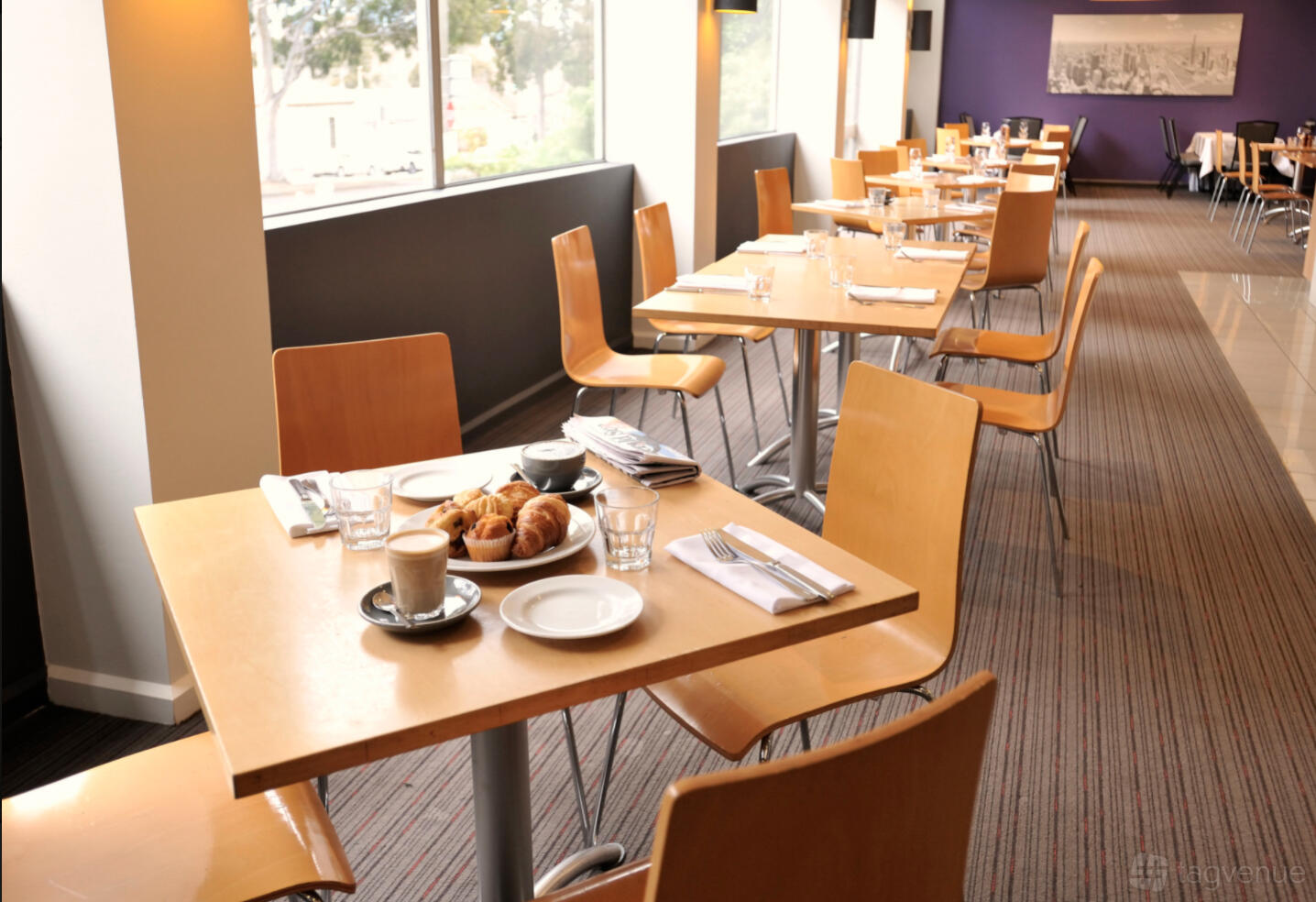 A cafe with wooden tables set with coffee, pastries, and cutlery by large windows at Mercure North Melbourne.