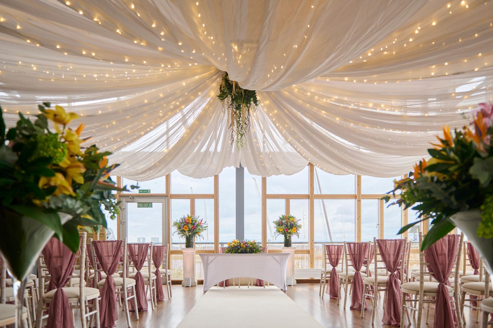An event venue with draped fabric ceiling, string lights, and large windows overlooking the river at Greenwich Yacht Club.