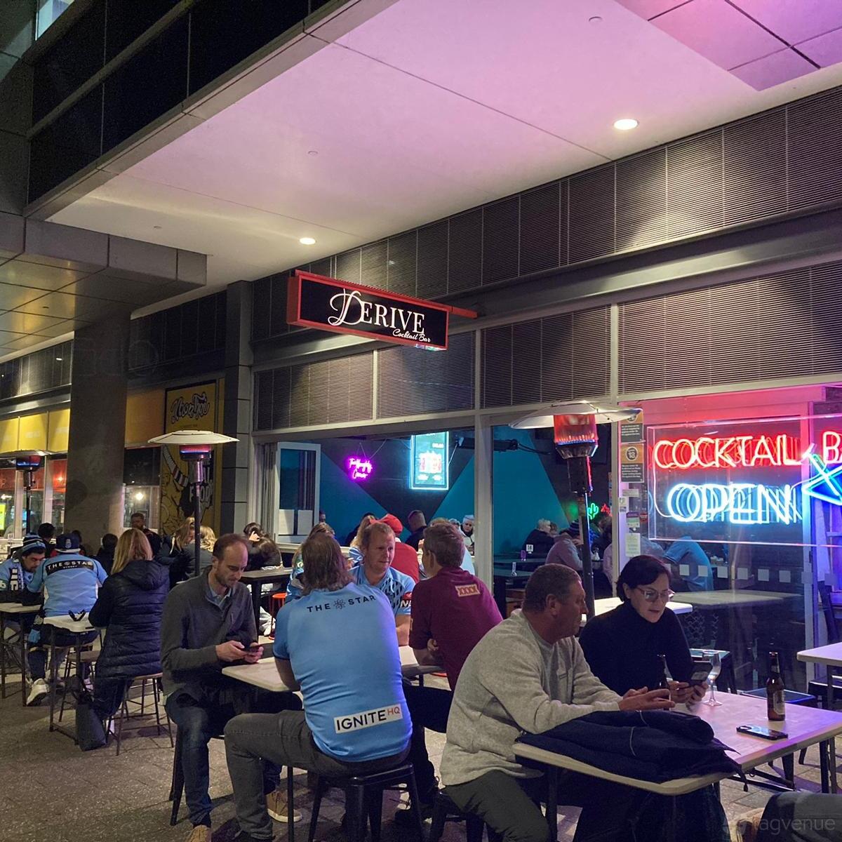 An outdoor bar area with neon signs, patio heaters, and groups seated at tables at Derive Cocktail Bar.