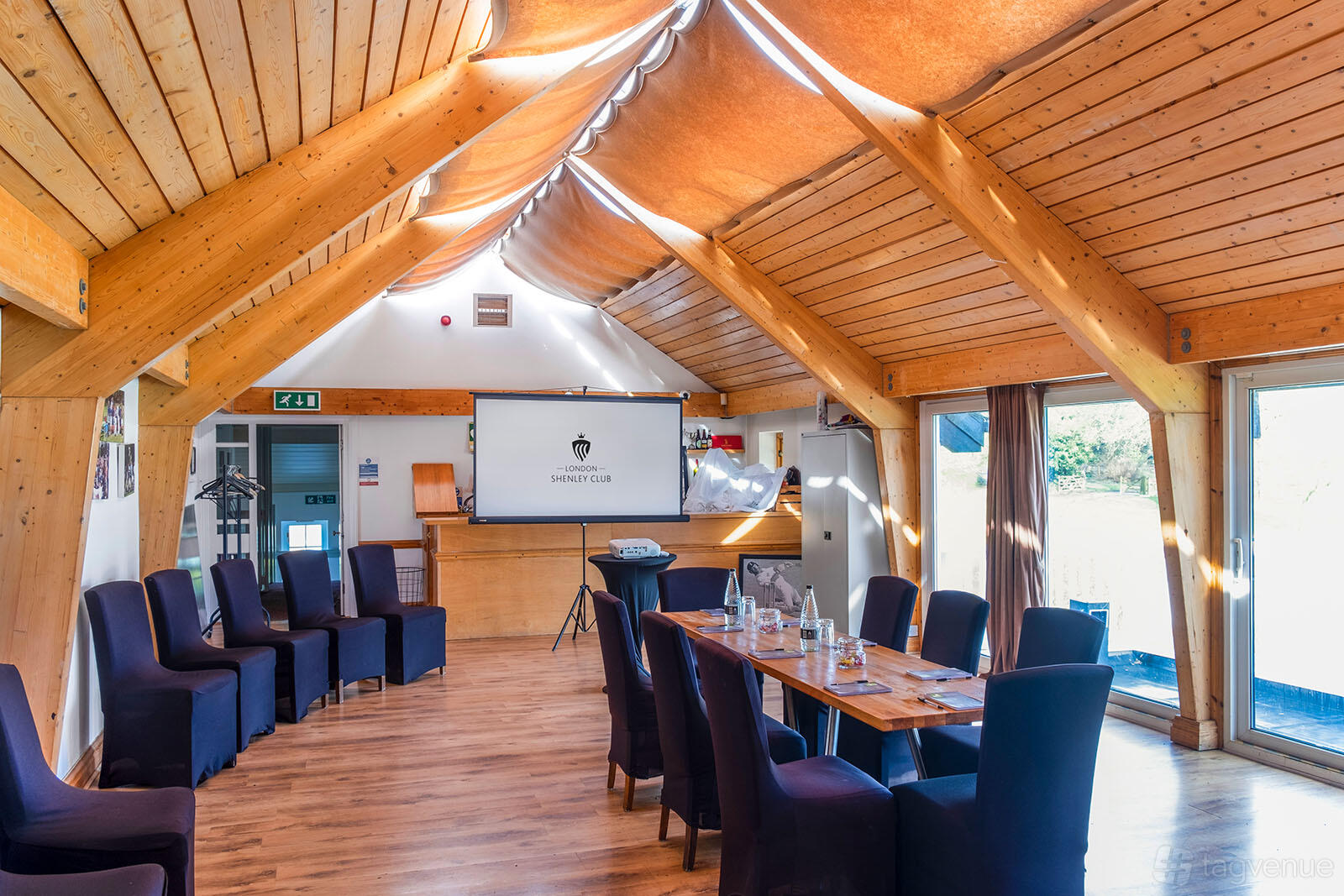 A meeting room with vaulted wooden ceilings, large windows, a long table with chairs, and a presentation screen at London Shenley Club.