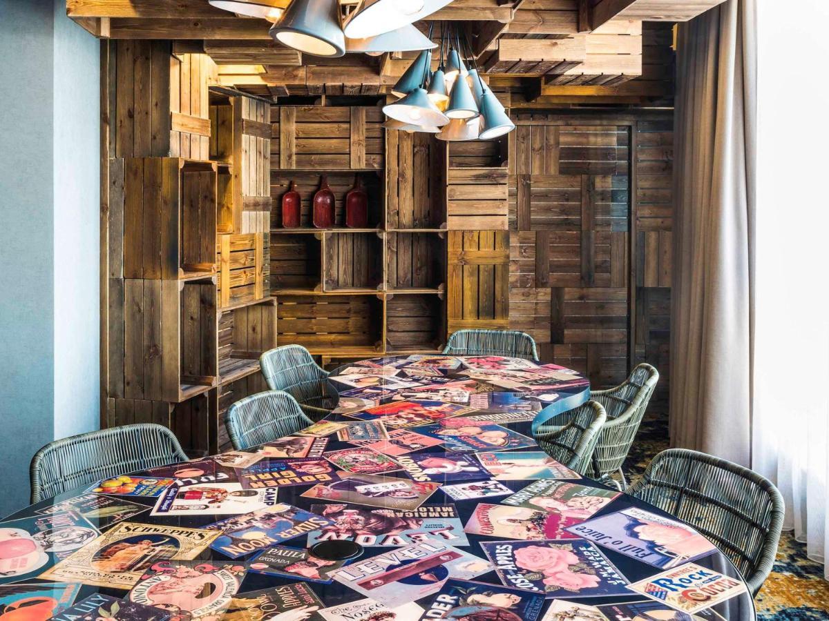 A meeting room with magazine-covered tables, wicker chairs, and wooden crate shelving at Novotel Canary Wharf.