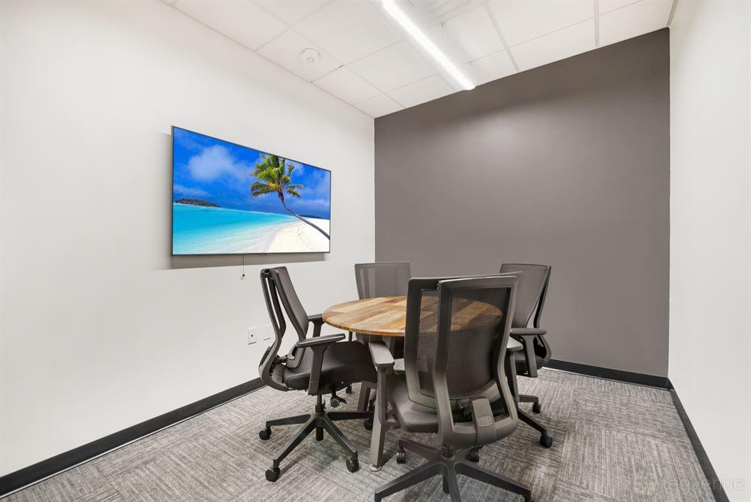 A meeting room with a round wooden table, four mesh chairs, and a large wall-mounted screen at Venture X Palmetto Bay.