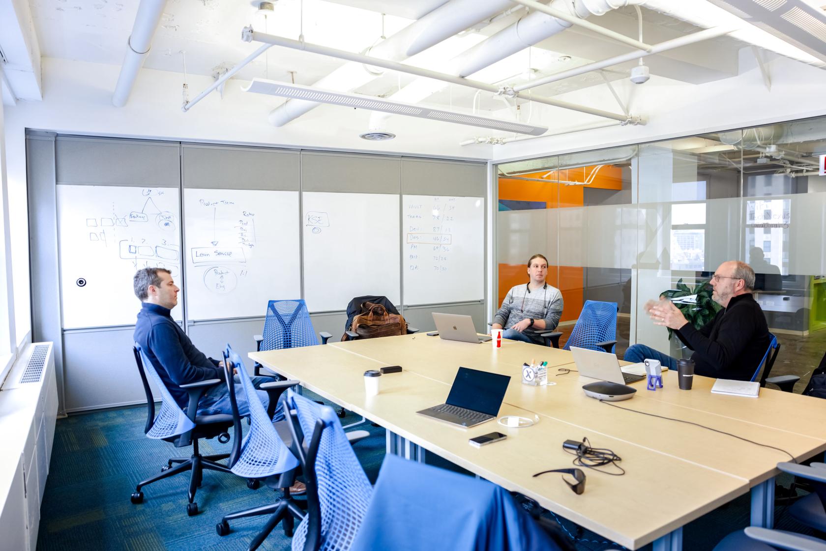 A boardroom with a long table, mesh chairs, large whiteboards, and exposed ductwork at TeamWorking by TechNexus.