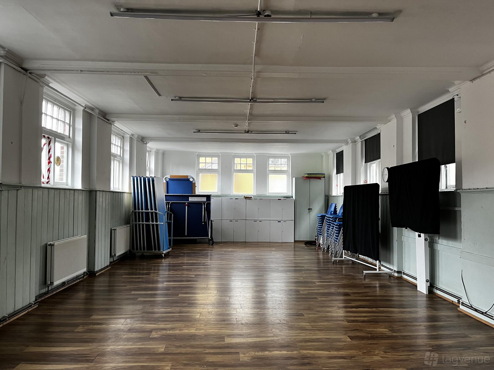 An event space with wooden flooring, stackable chairs, folding tables, and large windows at Katherine Low Settlement.