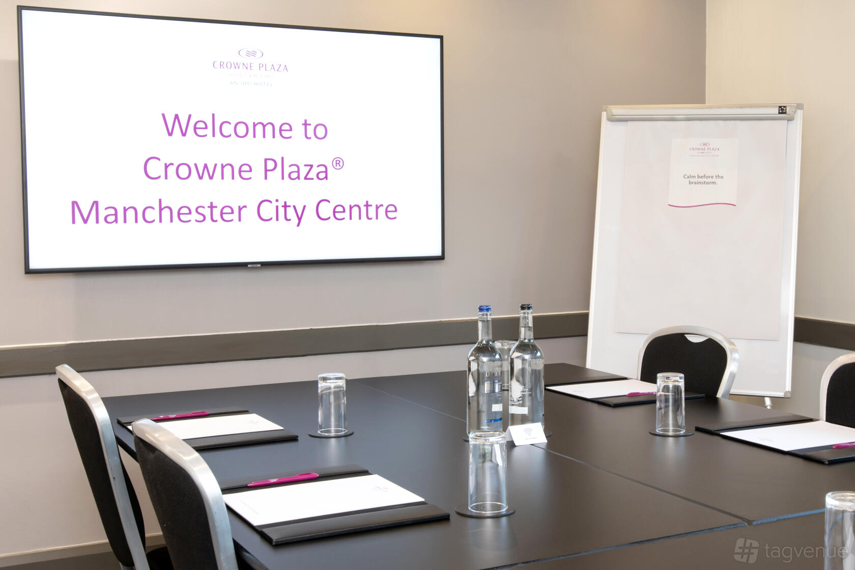 A hotel boardroom with a large display screen, flipchart, bottled water, and notepads at Crowne Plaza Manchester City Centre.