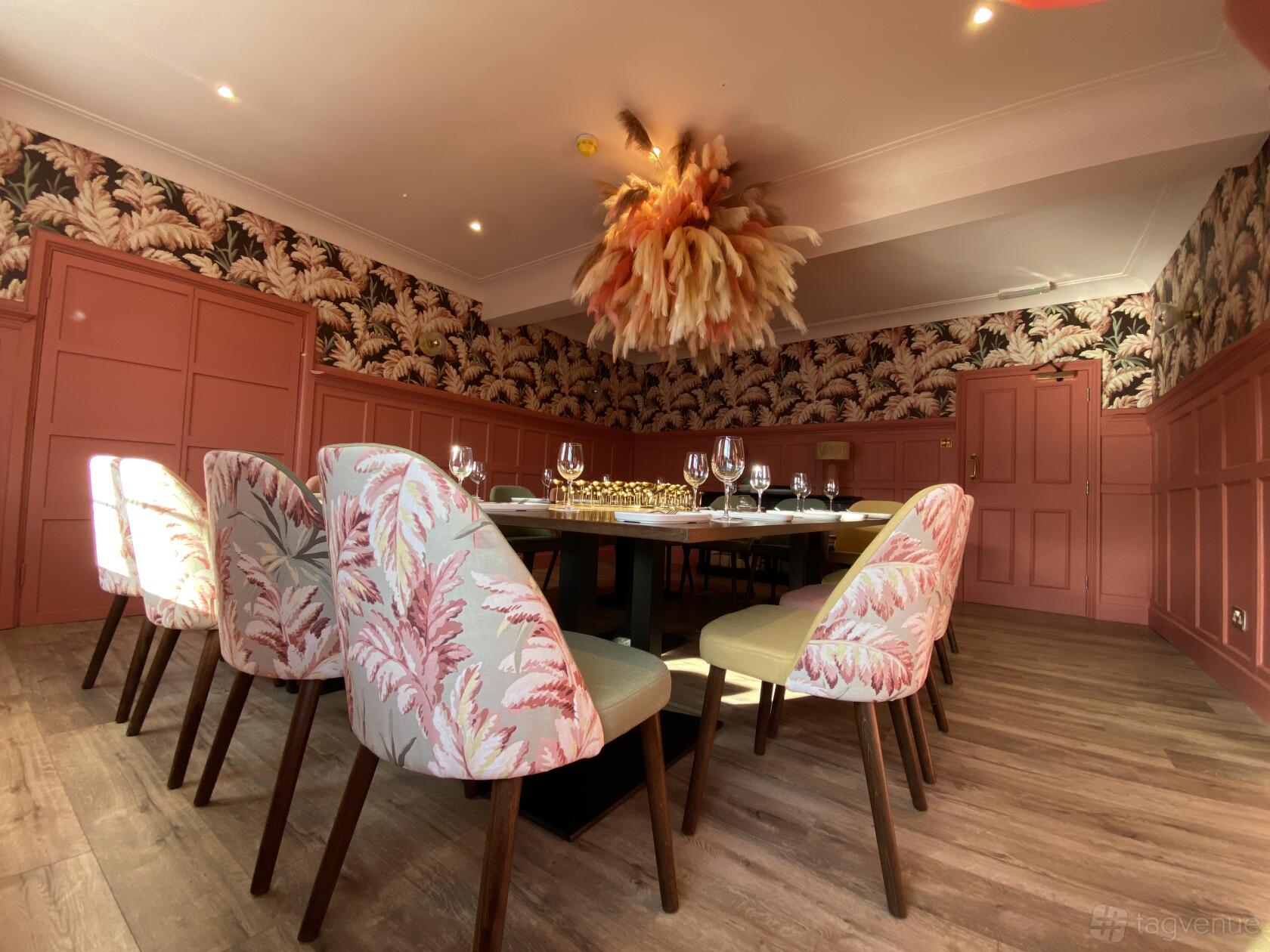 A dining room with patterned pink chairs, a large rectangular table, and a feathered chandelier at The Victoria.