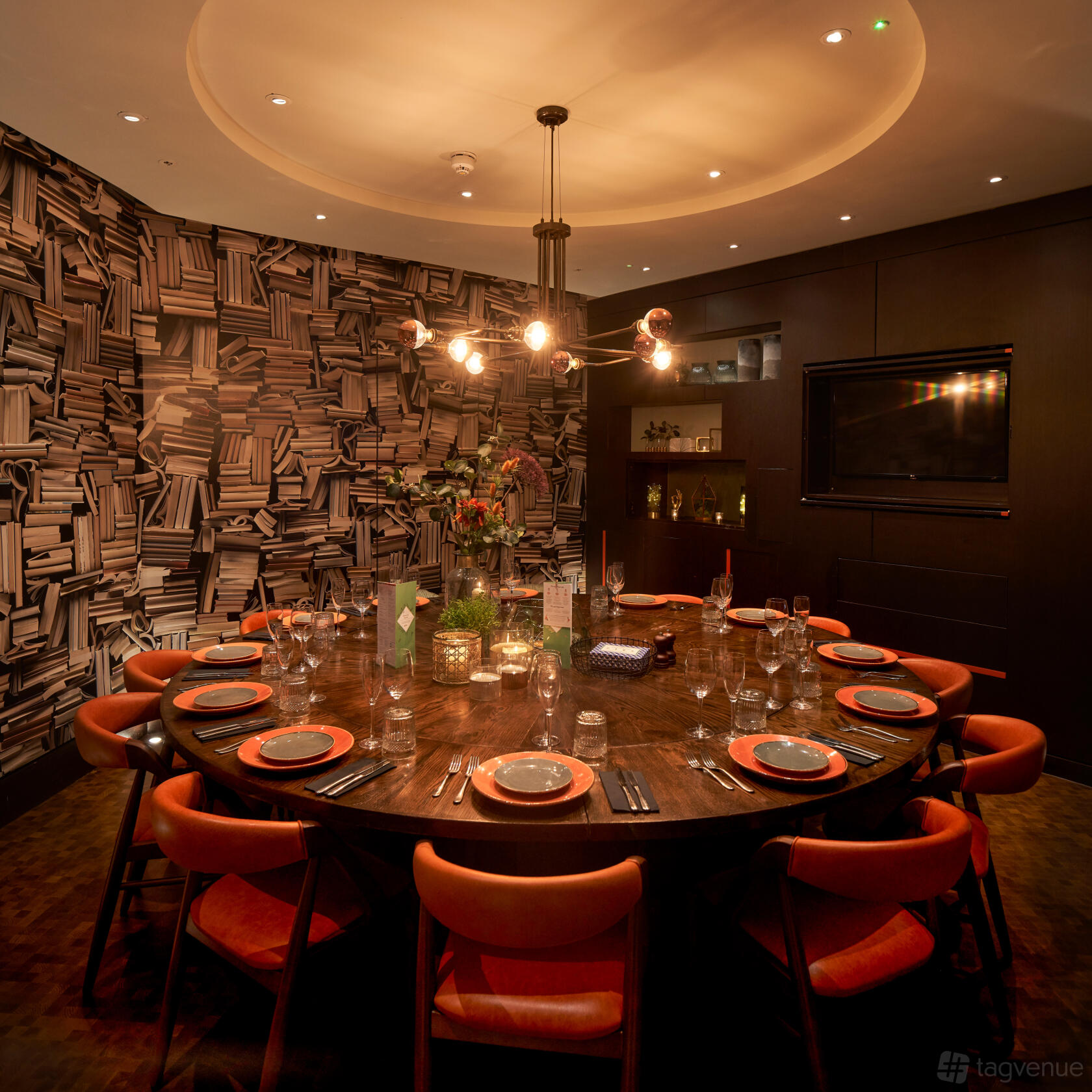 A private dining room with a round wooden table, orange chairs, and a bookshelf-patterned wall at The Moniker.