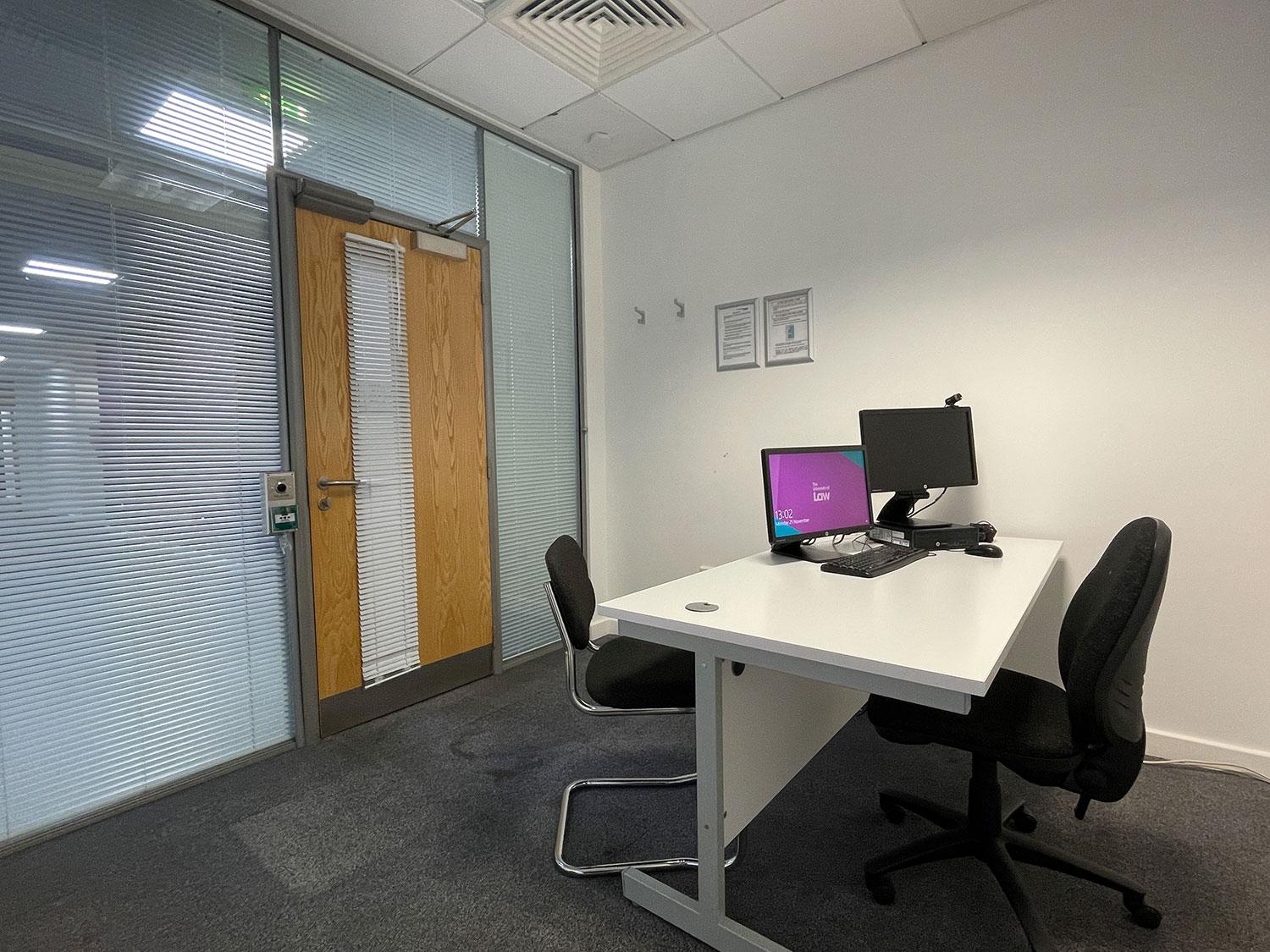 A meeting room with two office chairs, a white desk, desktop computer, and frosted glass wall at University of Law - London Moorgate.
