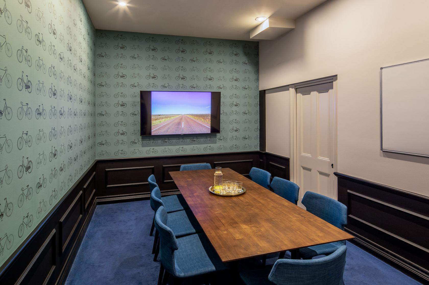 A meeting room with a wood conference table, blue chairs, wall-mounted screen, and patterned wallpaper at Christie Spaces Conferencing Melbourne.