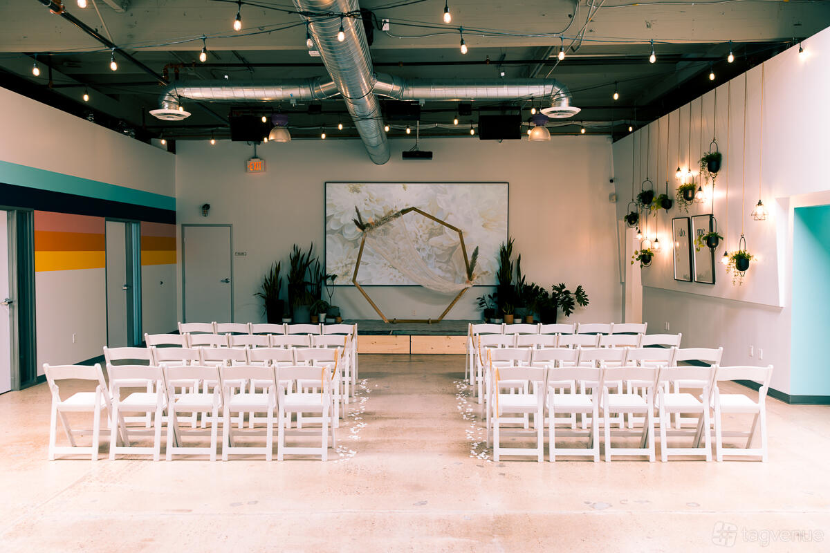 A warehouse venue with white chairs arranged for a ceremony, exposed ductwork, string lights, and potted plants at Rogue Collective.