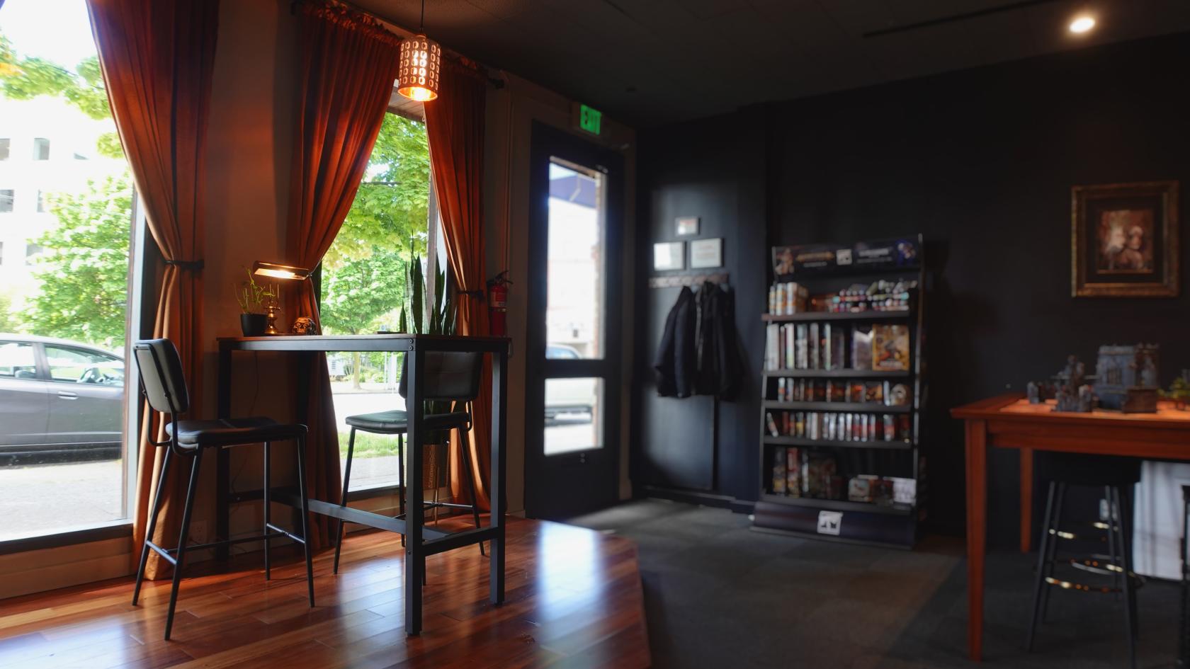 A lounge in a bar with wood floors, tall windows with orange curtains, and a bookshelf at Waystone Wargaming Speakeasy.