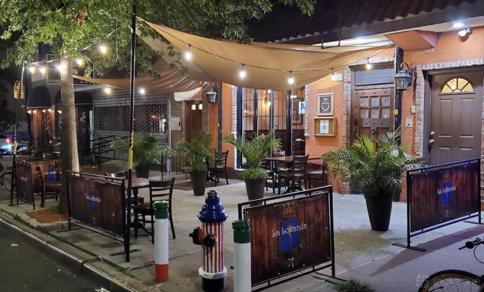 A restaurant terrace with string lights, potted plants, and shaded seating under canvas sails at La Locanda.