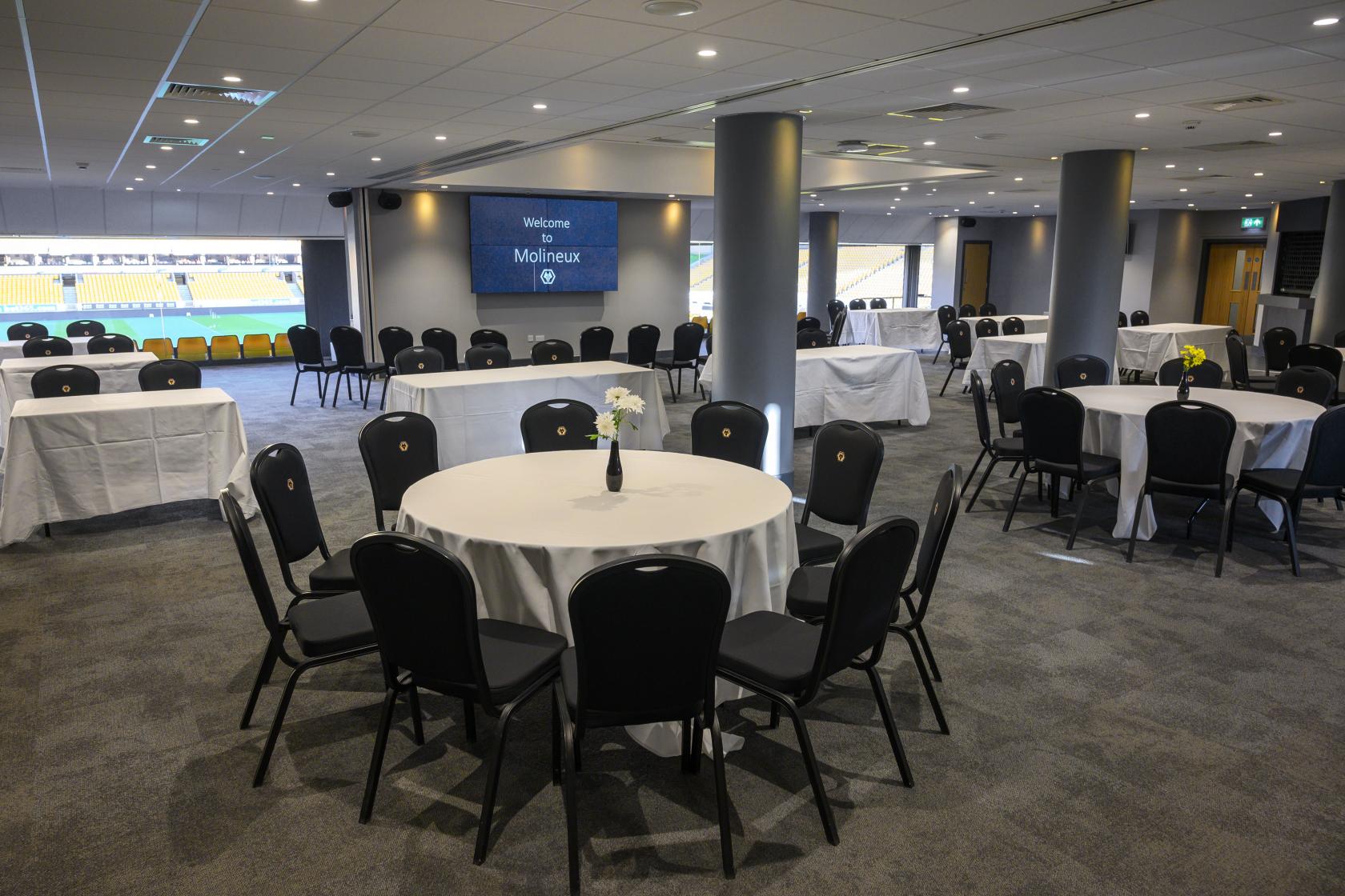 An event suite with round tables, black chairs, and floor-to-ceiling windows overlooking the pitch at Molineux Stadium.