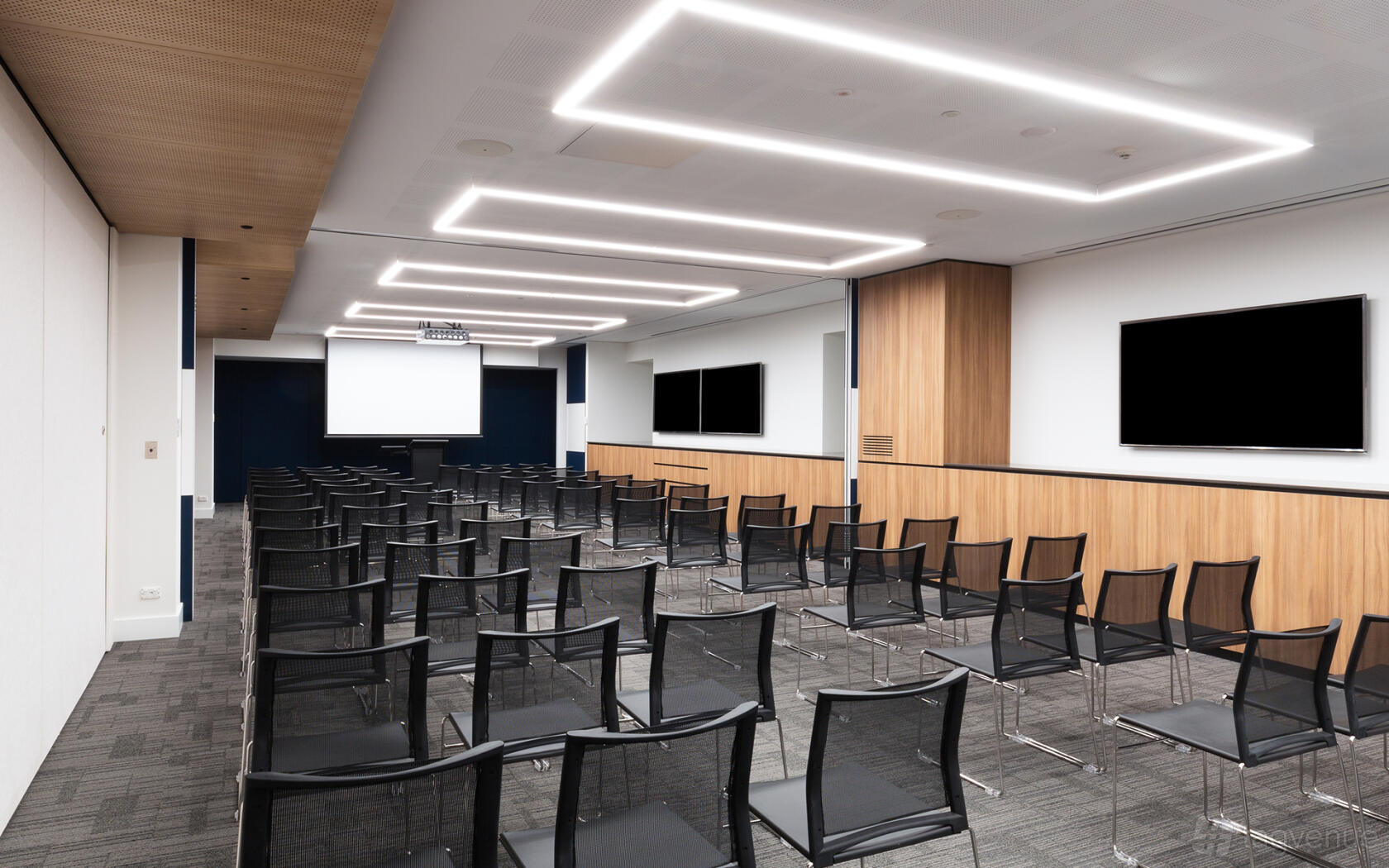 A meeting room with rows of black mesh chairs, wall-mounted screens, and a projector at Flex by ISPT at George Place.