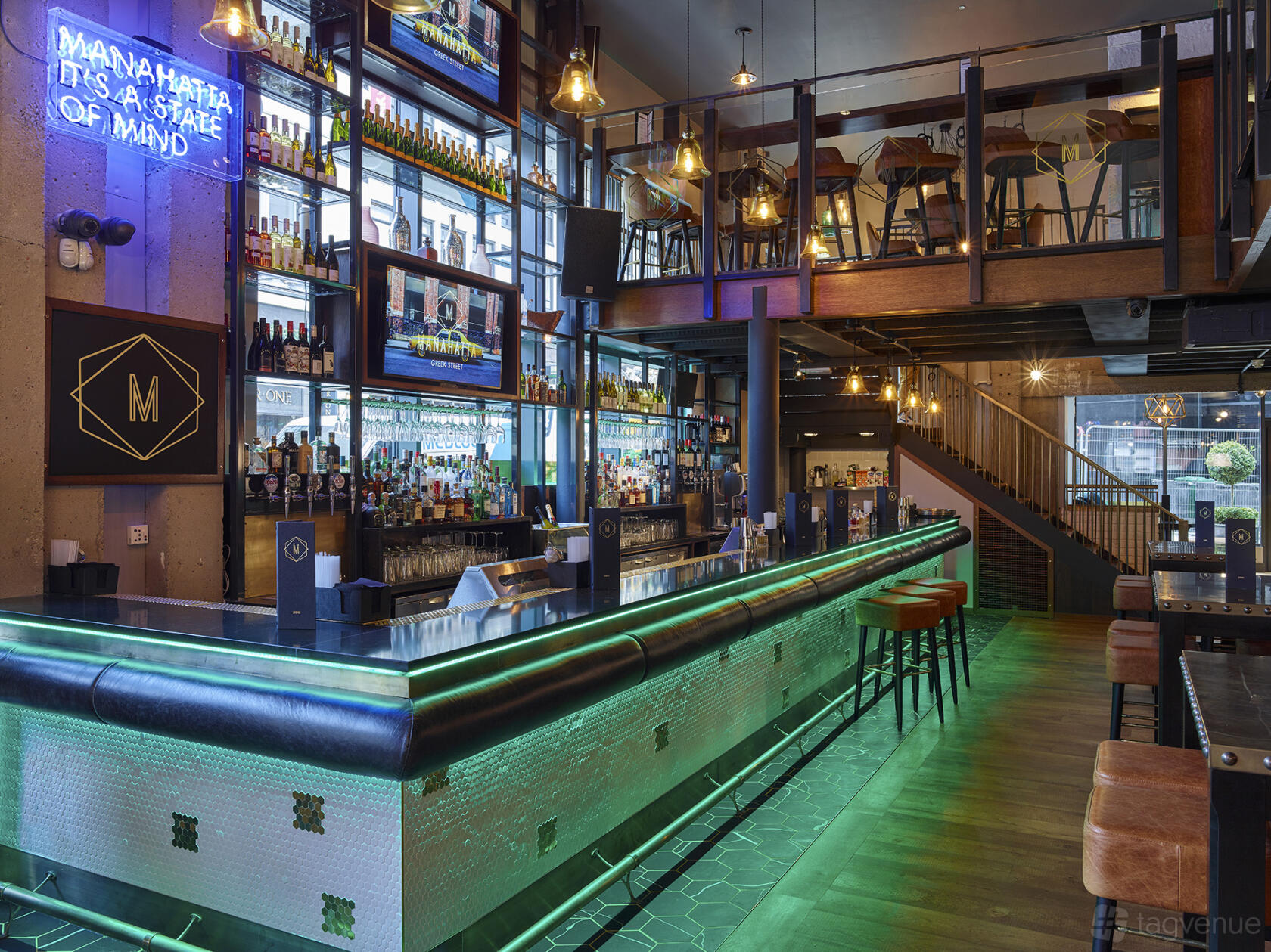 A cocktail bar with a long illuminated bar counter, hanging pendant lights, and a mezzanine at Manahatta Greek Street.
