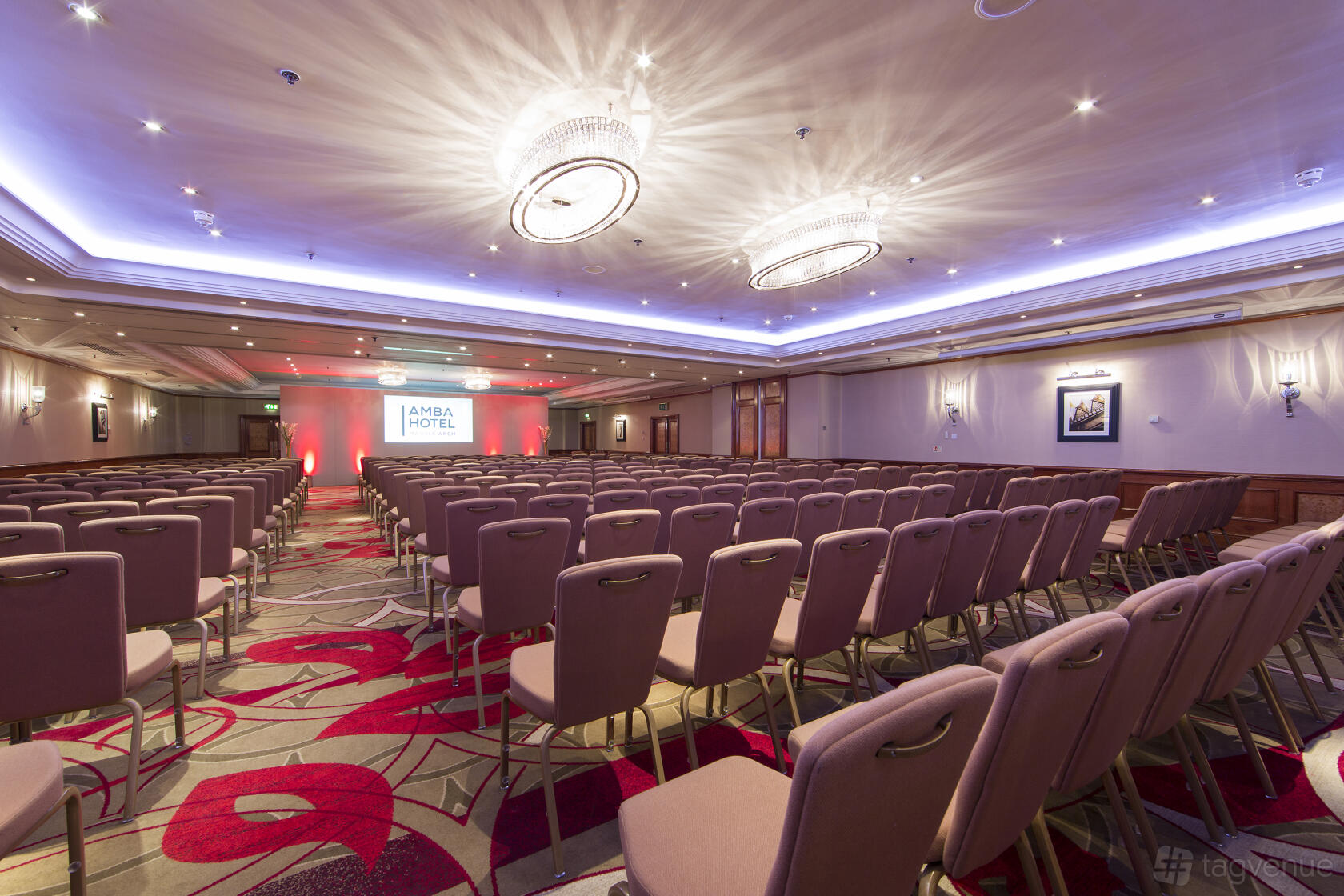 An auditorium with rows of upholstered chairs, patterned carpet, and ceiling chandeliers at The Marble Arch Hotel.
