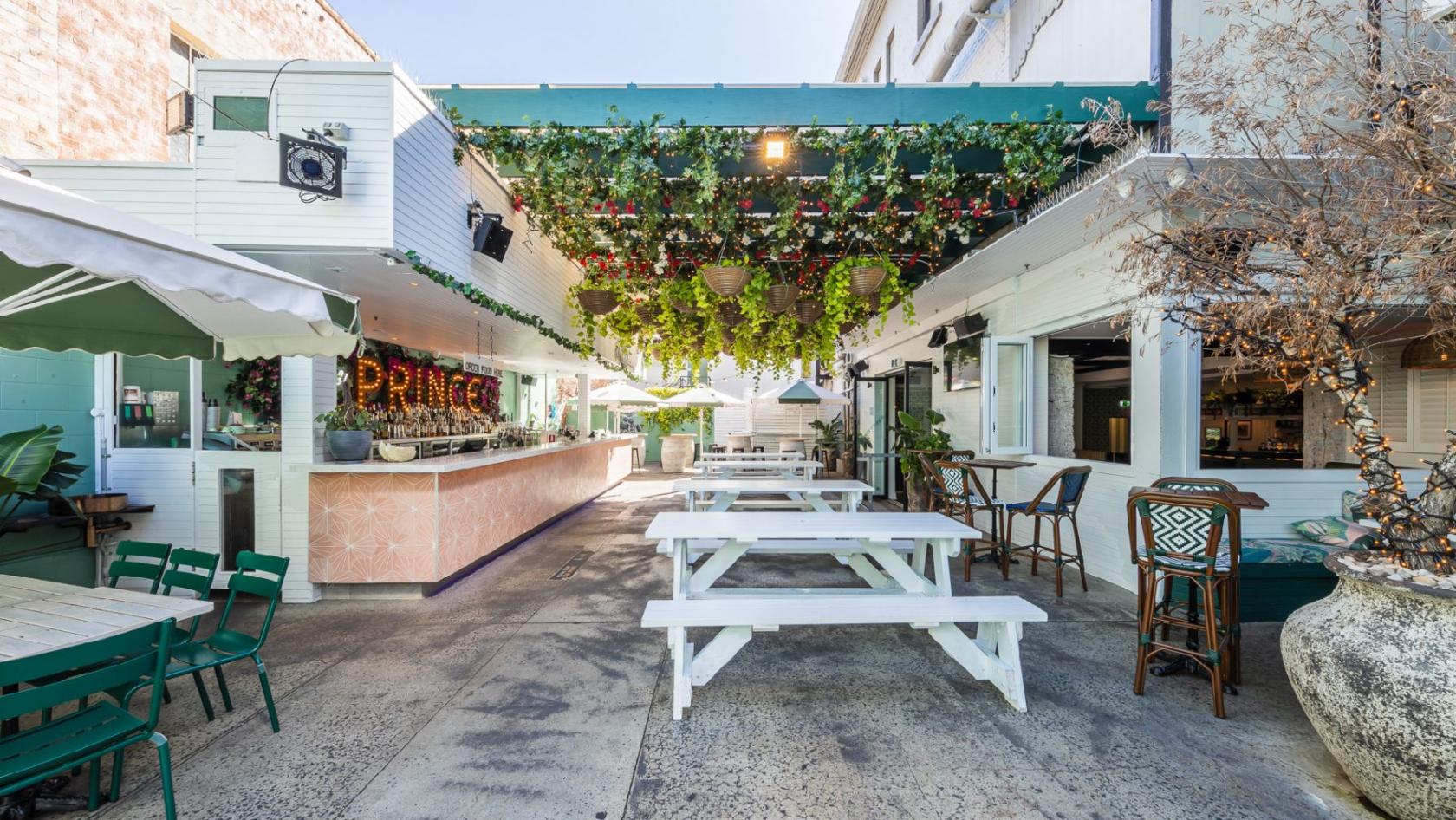 An outdoor pub area with white picnic tables, a vine-covered pergola, and a pink tile bar at The Prince Consort.