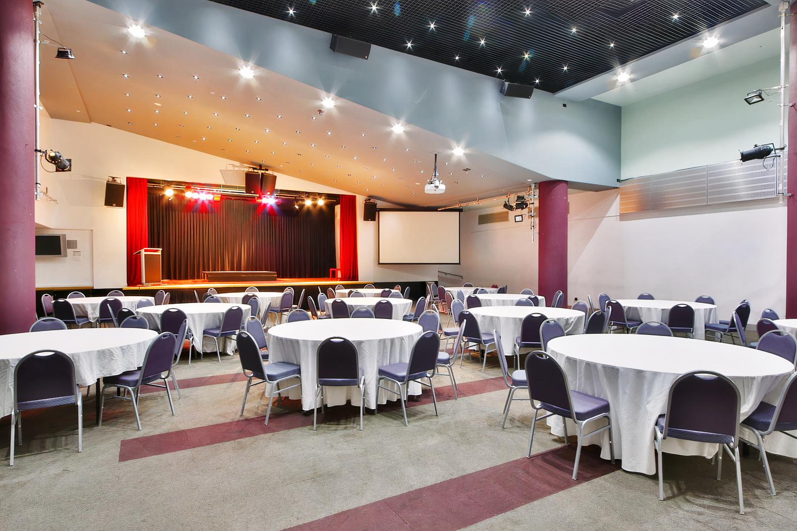 A hotel hall with round tables covered in white linens, stage with curtains, and ceiling spotlights at Arrow on Swanston.