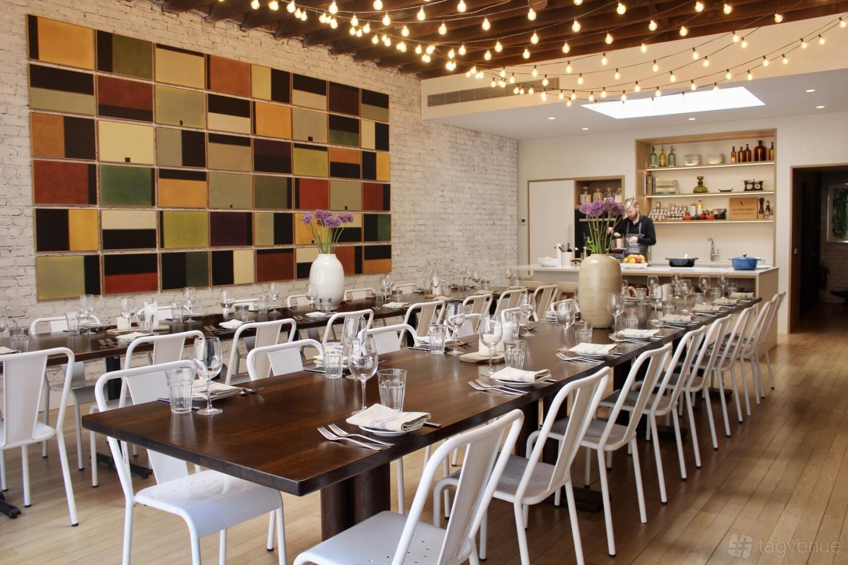 A kitchen with a long wooden dining table, white chairs, string lights, and open shelving at MM Kitchen Studio.