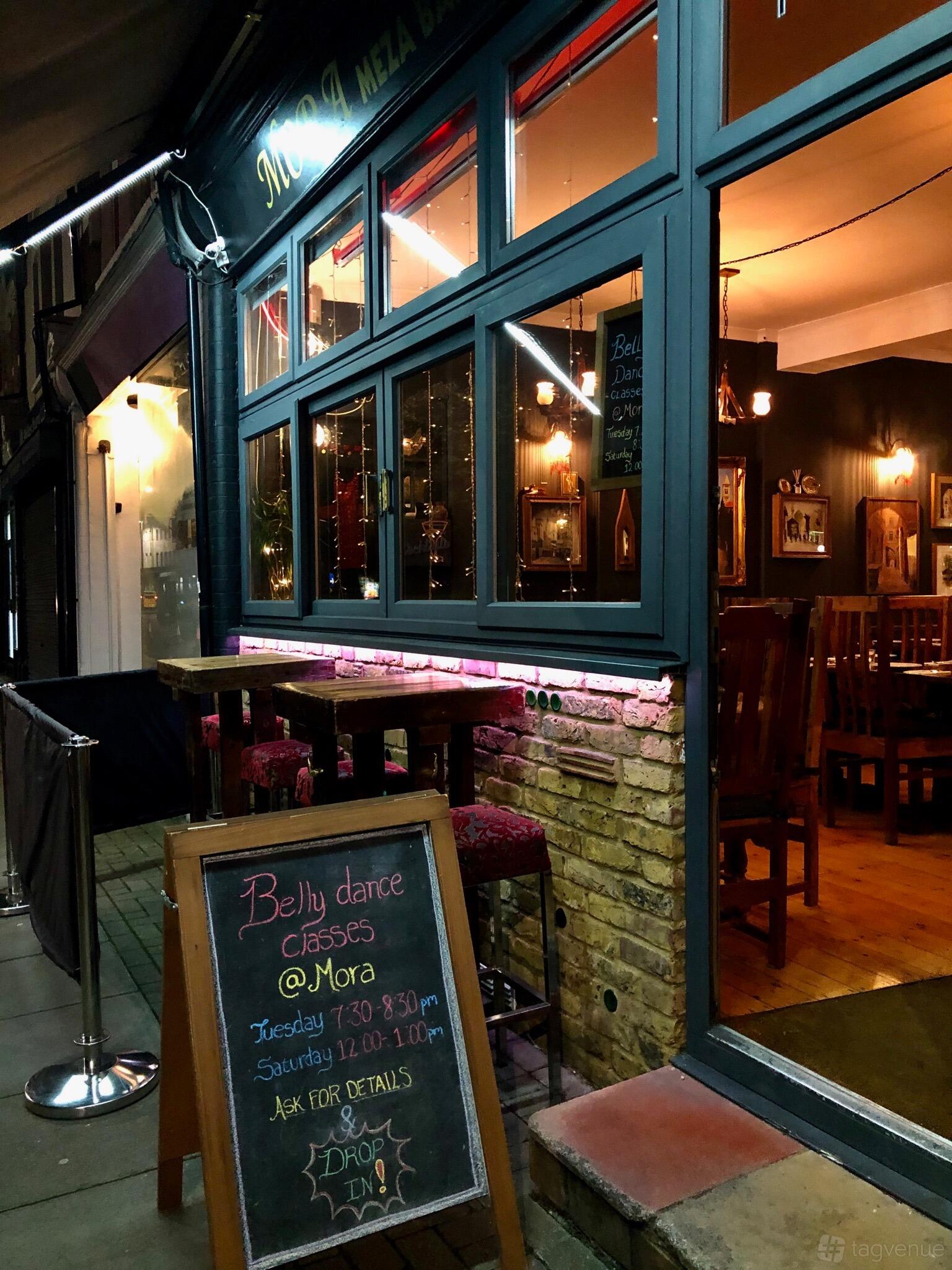 A restaurant exterior with brick walls, large windows, and outdoor wooden tables with upholstered stools at Mora Meza Bar & Restaurant.
