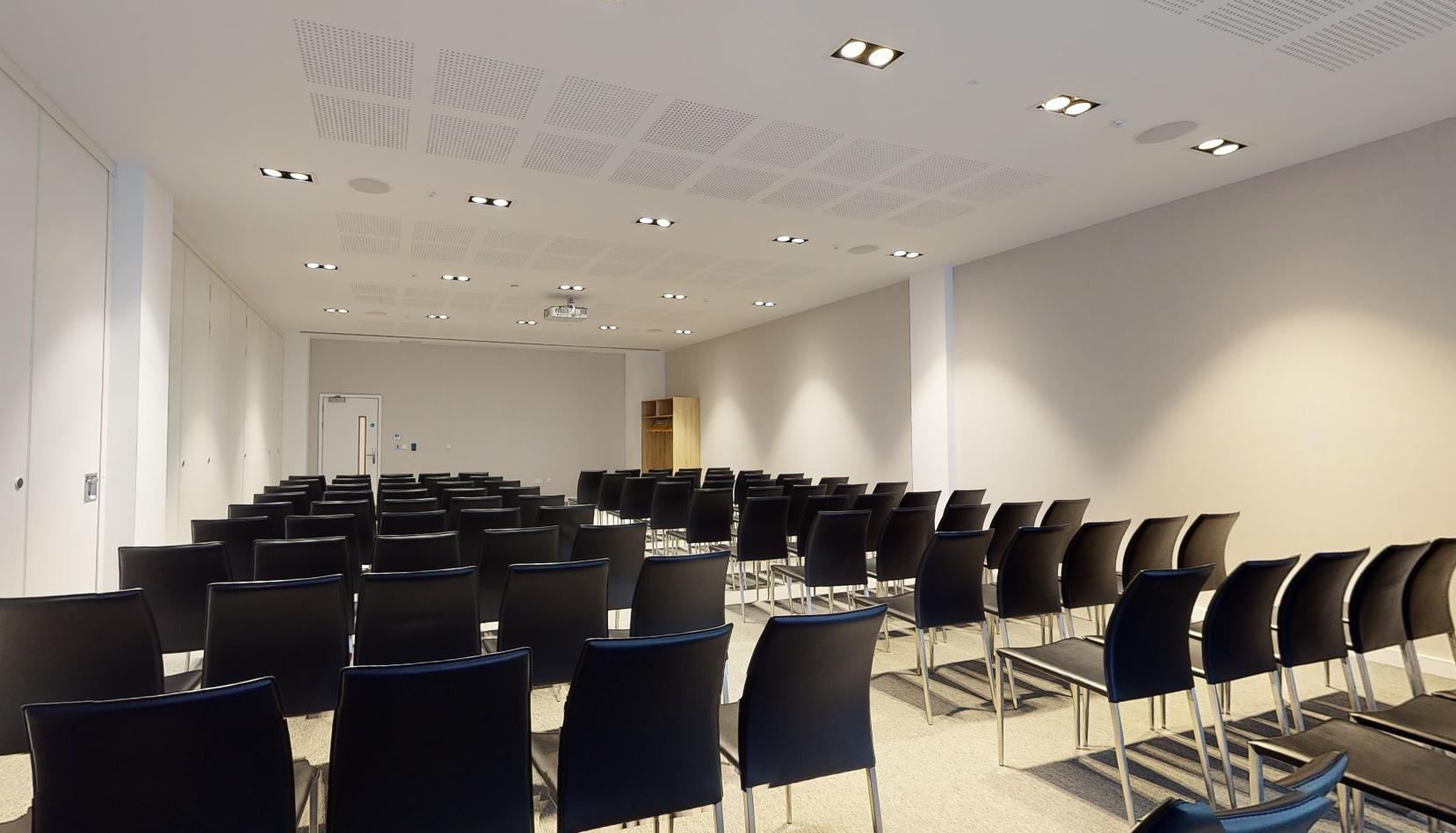 A hotel meeting room with rows of black chairs, ceiling lights, and a projector at INNSiDE by Melia Liverpool.
