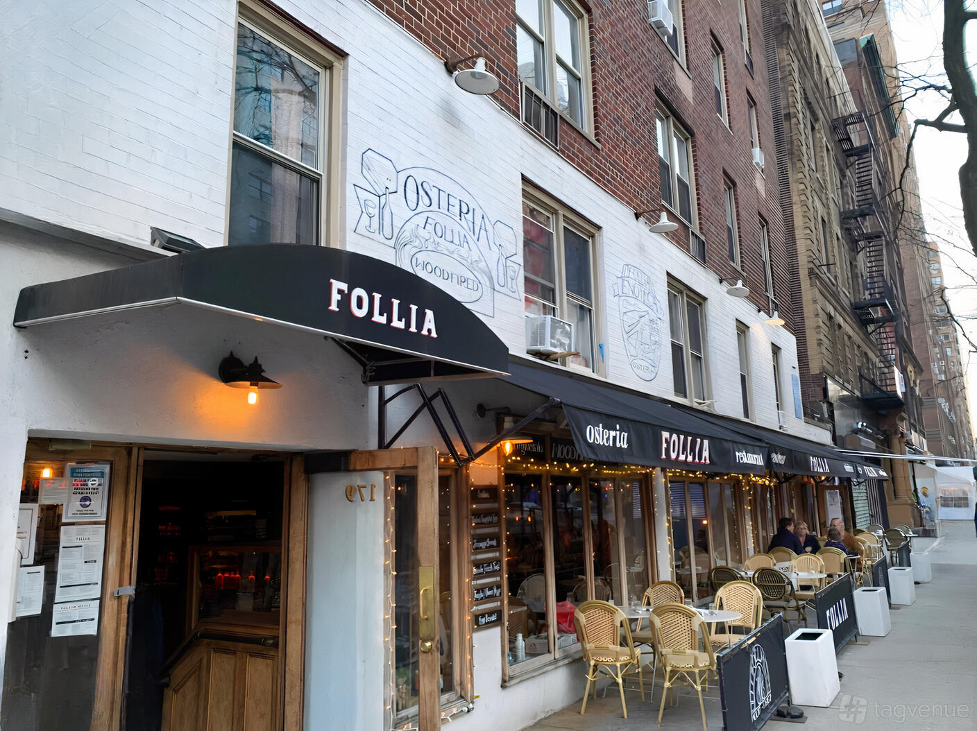 A restaurant with outdoor seating, black awnings, and string lights along a city sidewalk at Follia.