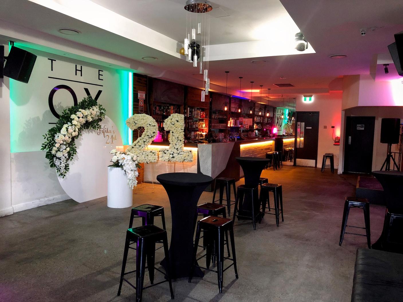 A bar with a long illuminated counter, high tables with black stools, and floral number decorations at The Ox Darlinghurst.