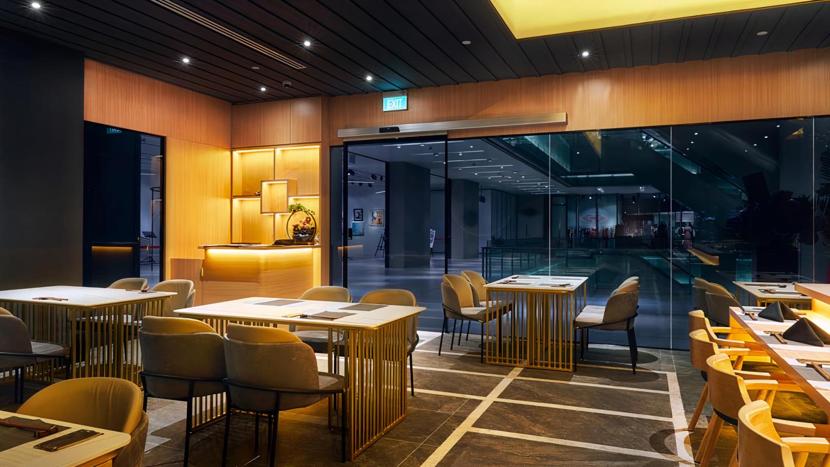 A restaurant with gold metal tables, cushioned chairs, wood paneling, and floor-to-ceiling glass doors at Kakushin.