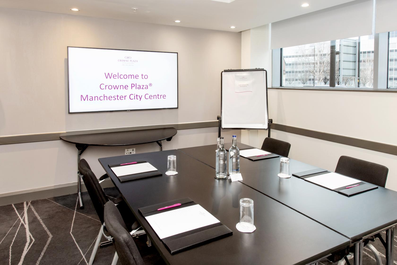 A hotel boardroom with a rectangular table set with notepads, water bottles, glassware, and a screen at Crowne Plaza Manchester City Centre.