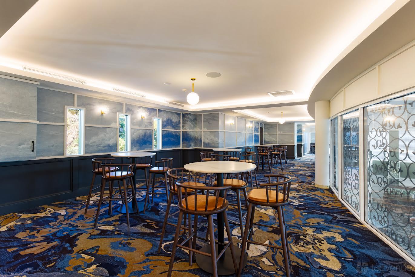 A pub event room with patterned carpet, round tables with wooden chairs, and large windows at Rocksia Hotel.