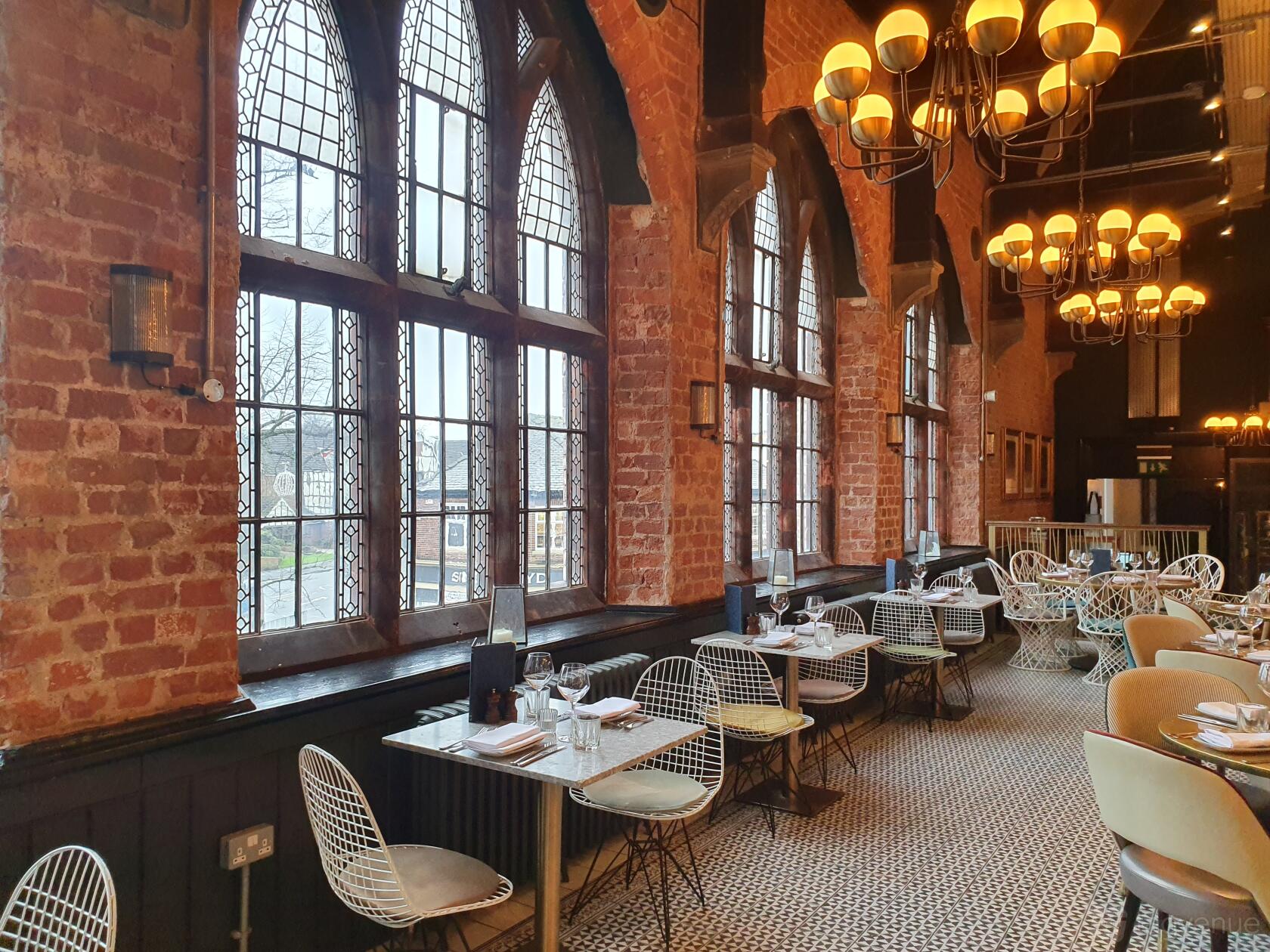 A restaurant bar with exposed brick walls, large arched windows, and chandeliers at The Lost and Found Knutsford.