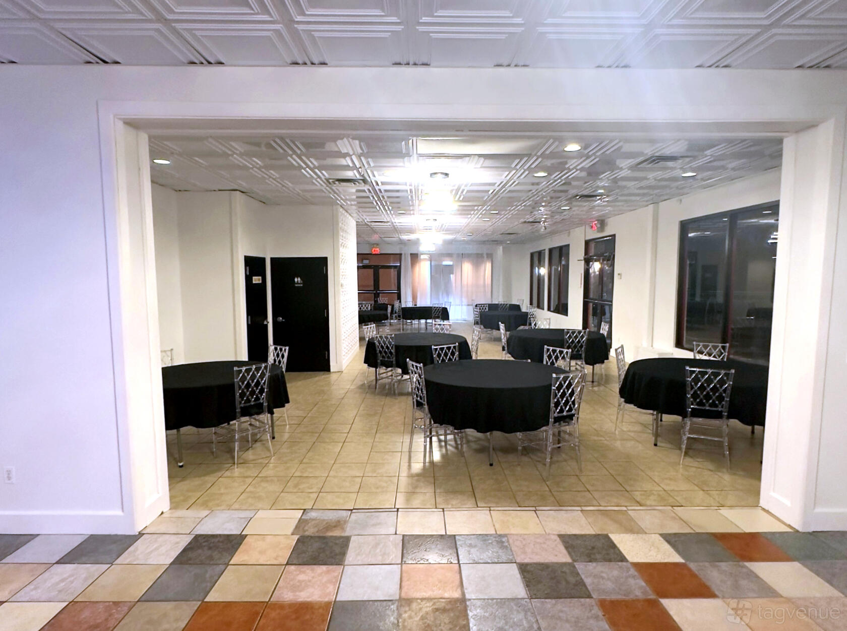 An event venue with round tables draped in black linens and clear chairs on a tiled floor at The Riviera Room.