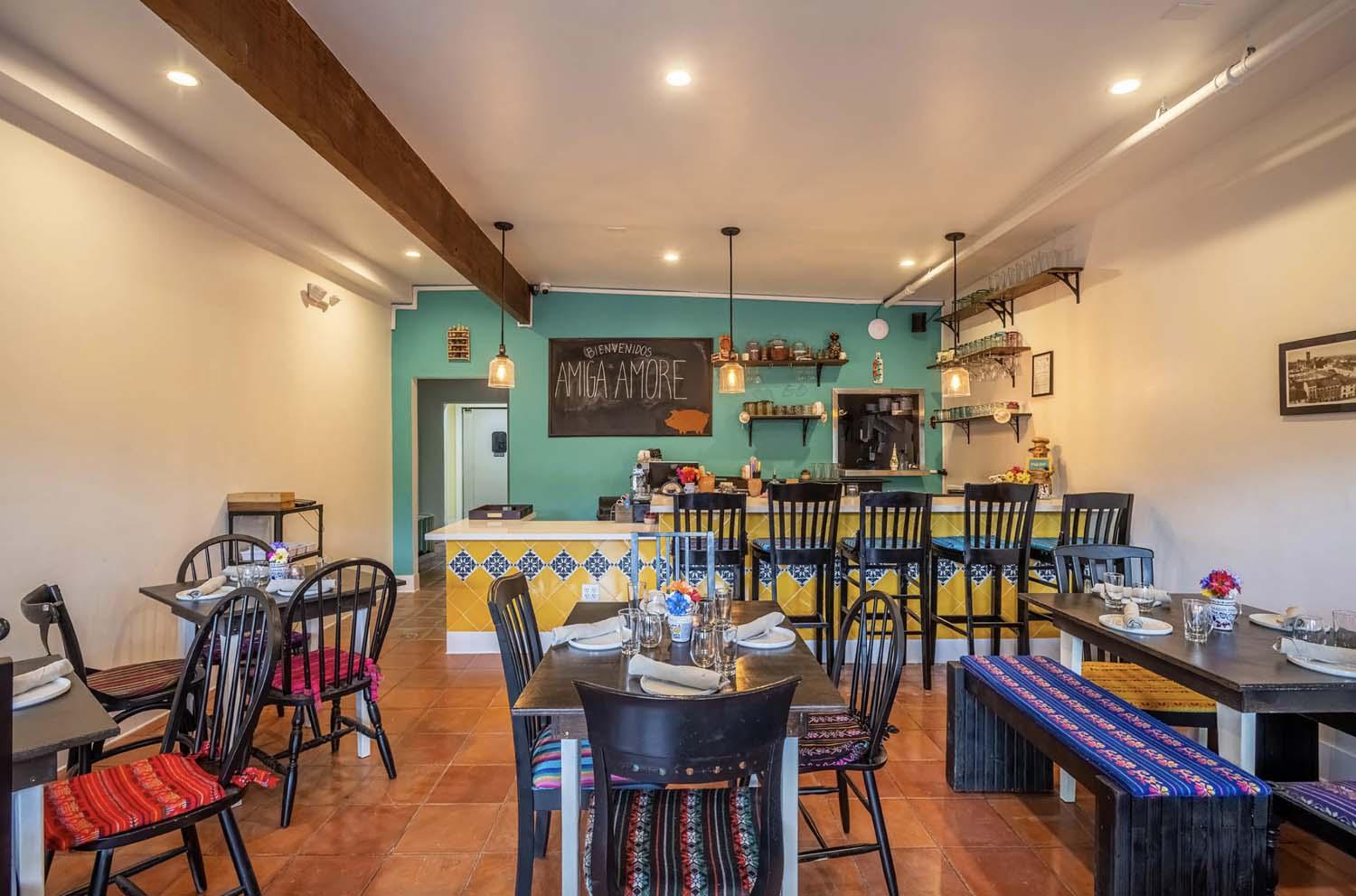 An event space in a restaurant with colorful tiled bar, wooden tables, multicolored seat cushions, and green accent wall at Amiga Amore.