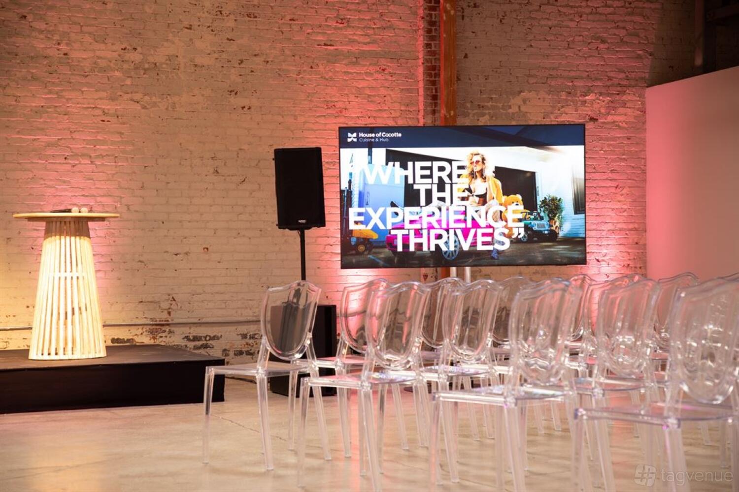 An event hall with exposed brick walls, clear acrylic chairs, and a large screen at House of Cocotte.