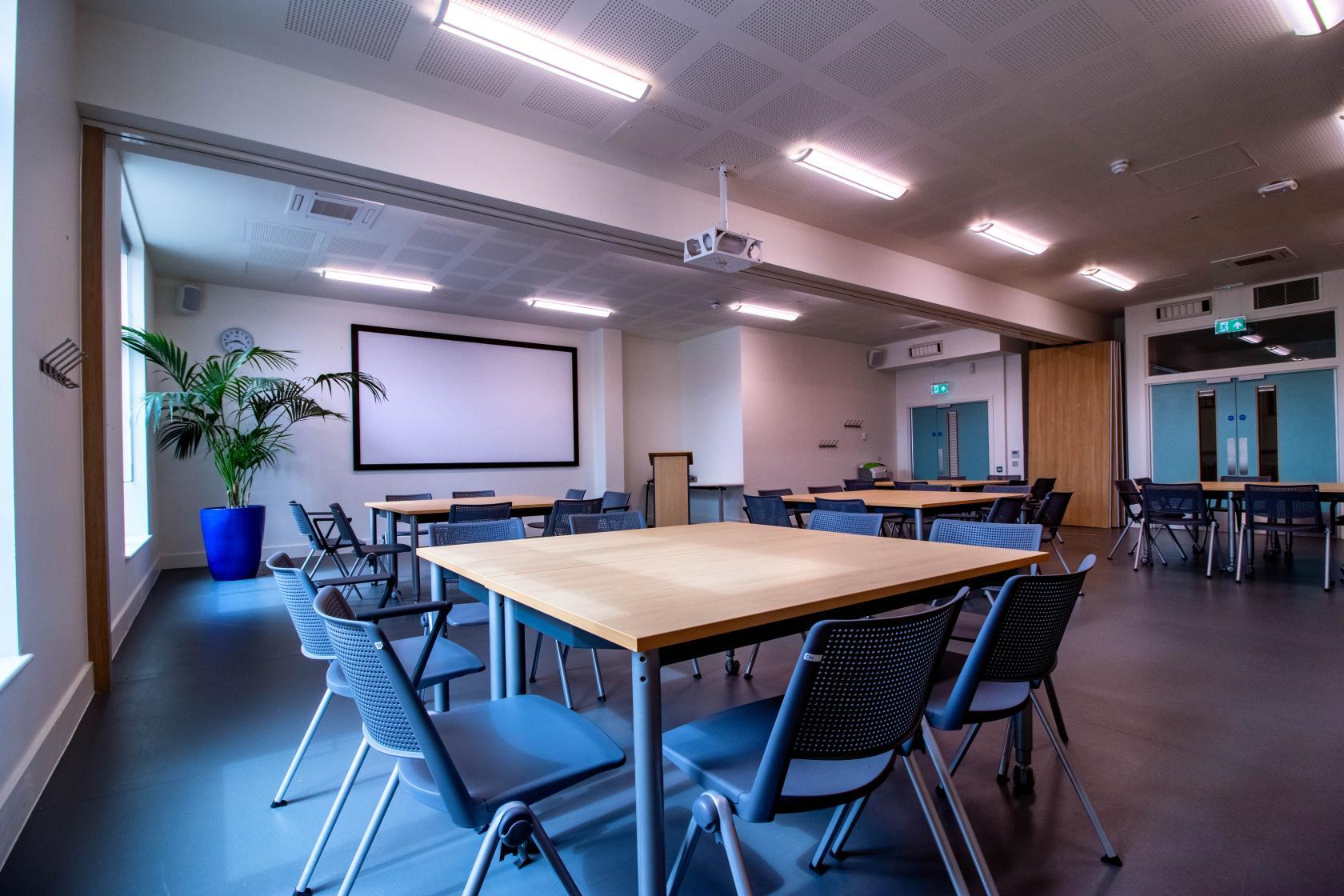 A hall with large windows, a projector screen, modern lighting, and neatly arranged tables at The Greenwood Centre.