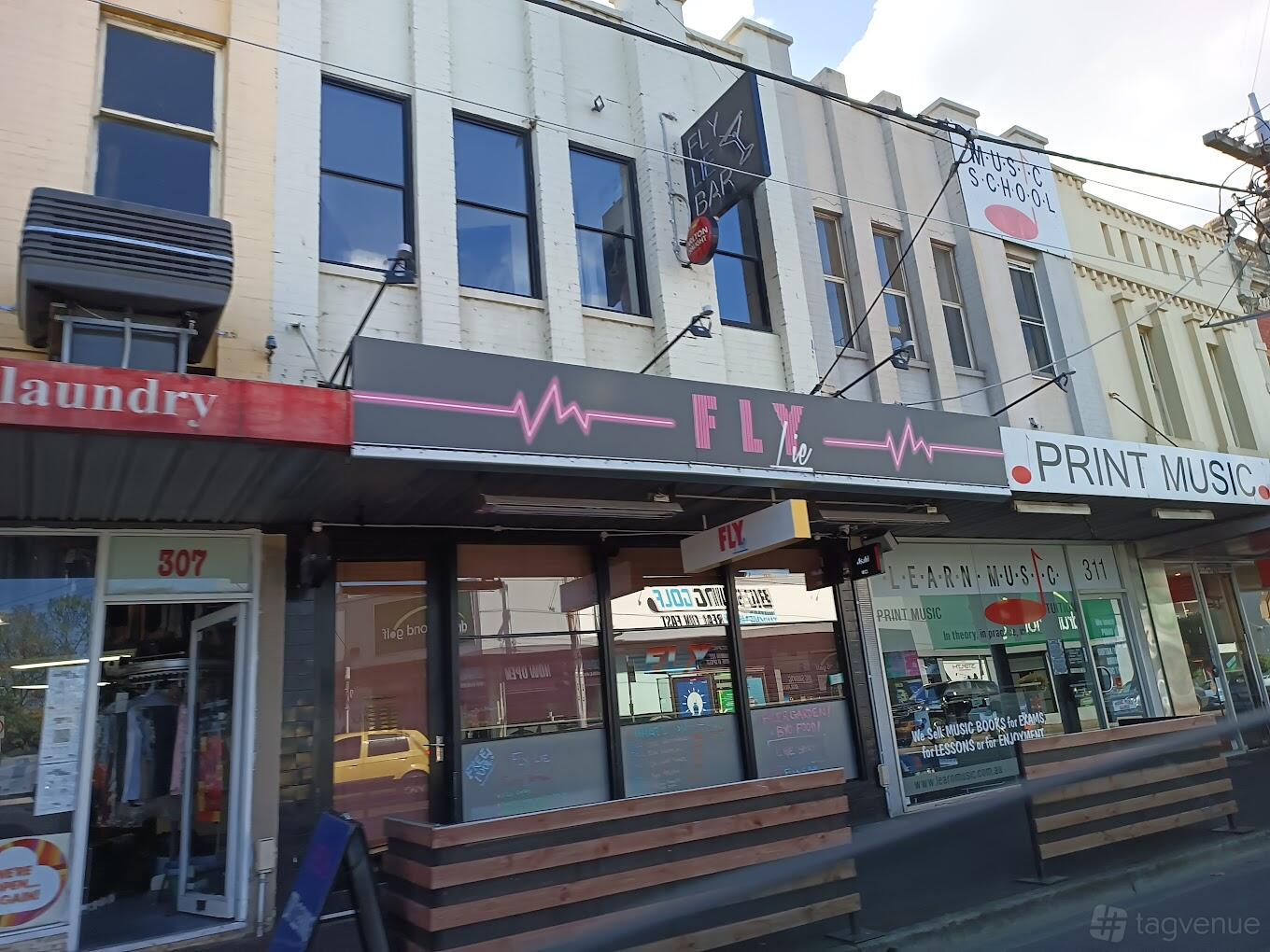 A pub with a dark exterior facade, black and pink neon signage, and large front windows at Fly Lie.
