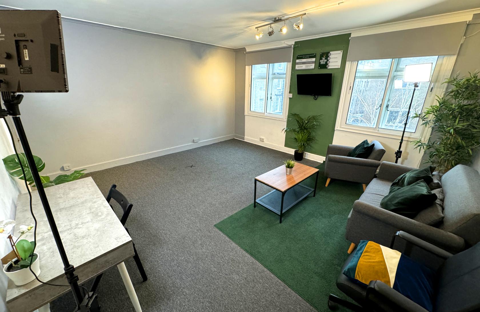 A rehearsal studio with large windows, a green accent wall, sofas, a desk, and potted plants at The Audition House 3x studios.