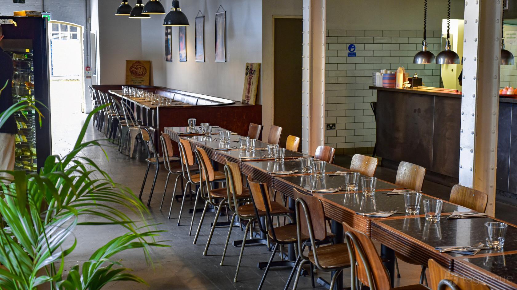 A dining room with long wooden tables, mismatched chairs, pendant lighting, and white tile walls at The Printhouse at Jim & Tonic East.