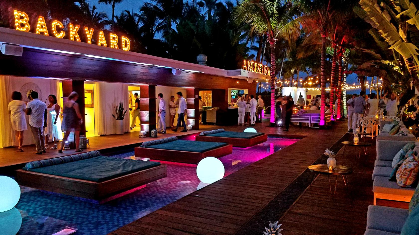 An outdoor hotel space with a pool, lounge beds, glowing orbs, string lights, and palm trees at The Marseilles Hotel.