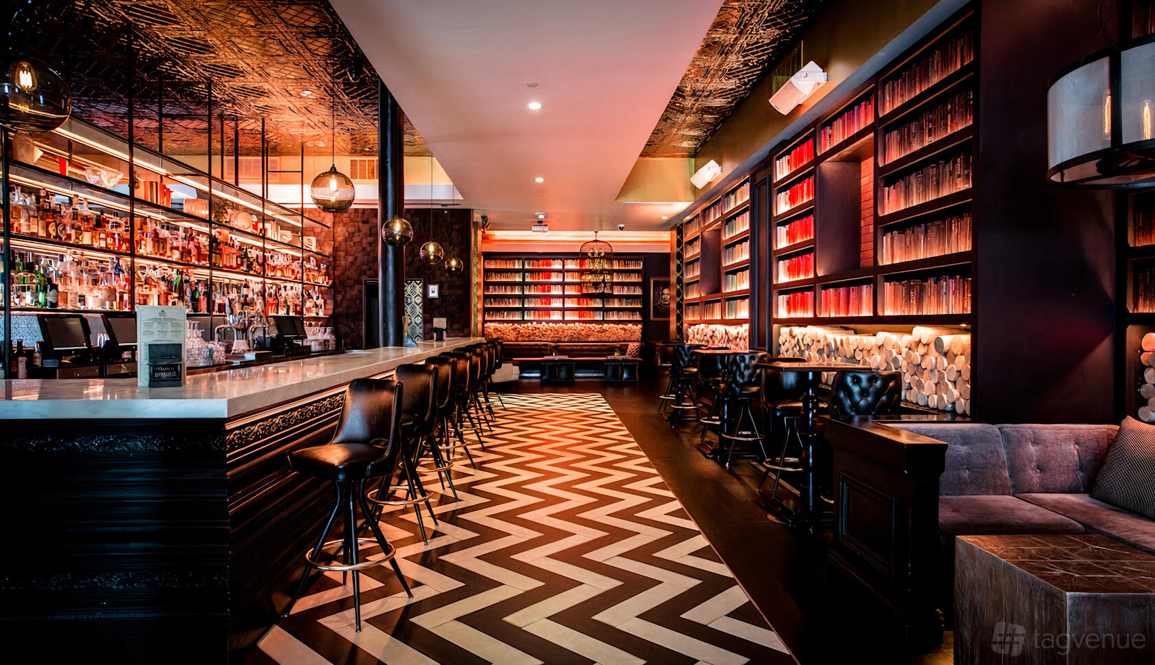 A cocktail bar with a marble-topped counter, chevron-patterned floor, backlit bookshelves, and leather bar stools at Novela.