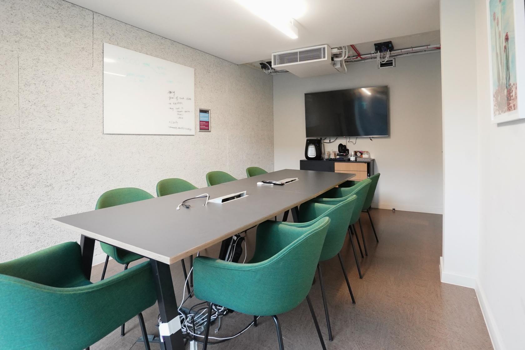A meeting room with green chairs, a long table, wall-mounted screen, and a whiteboard at The London Interdisciplinary School.